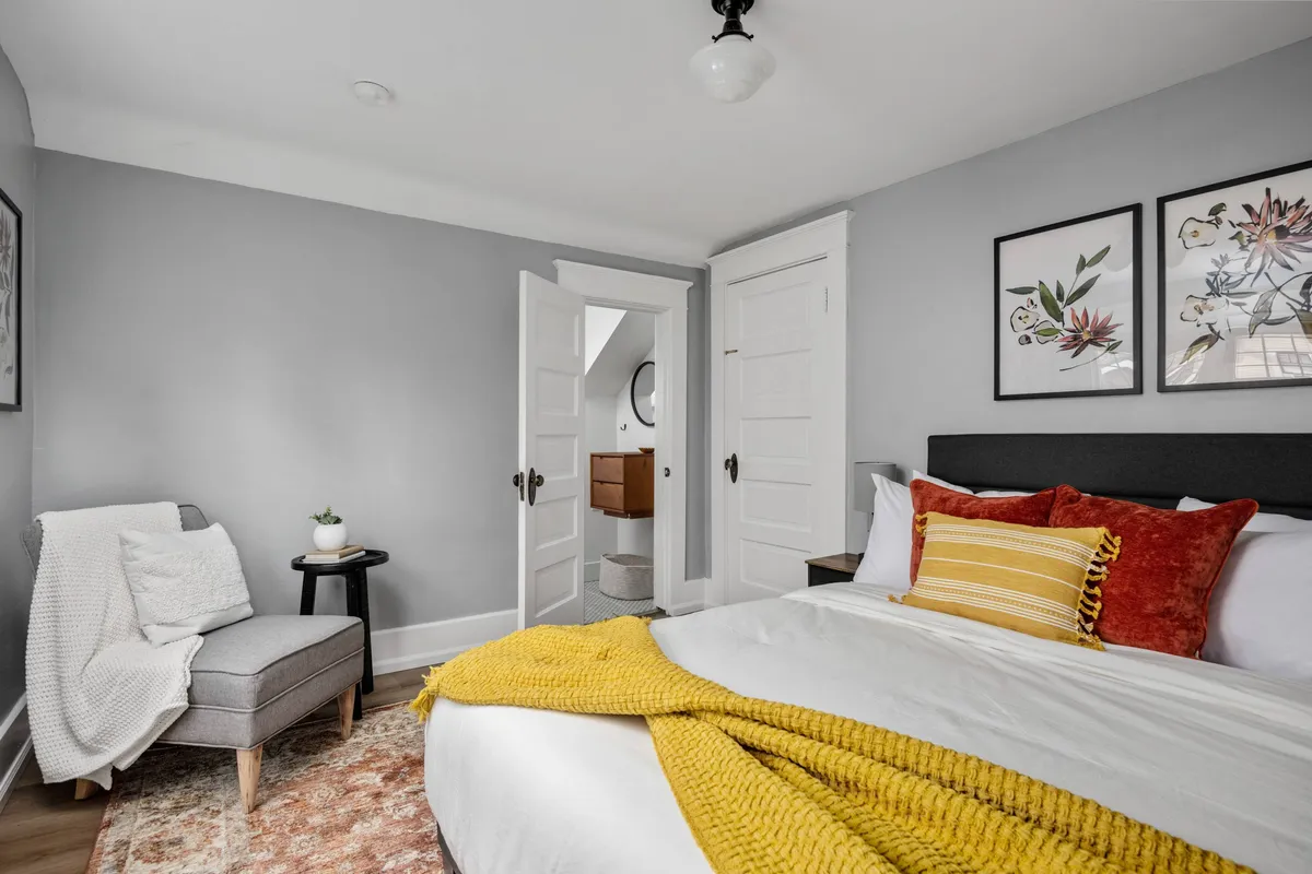 A tranquil bedroom scene featuring a neatly made bed with a sunny yellow throw blanket, tasteful artwork on the wall, and an inviting armchair draped with a cozy blanket. The space is bright, with natural light streaming in through the window, highlighting a peaceful atmosphere perfect for unwinding or curling up with a book.