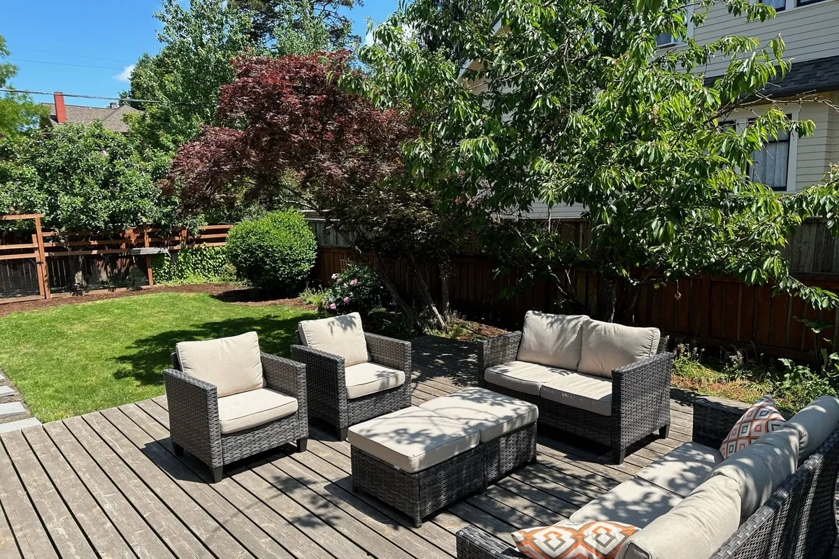 A tranquil exterior scene showcasing a spacious backyard with a wooden deck, inviting outdoor seating with plush decorative cushions, and lush greenery all around. The setup suggests a space perfect for relaxation or gatherings, with the comfort of the seating drawing attention.