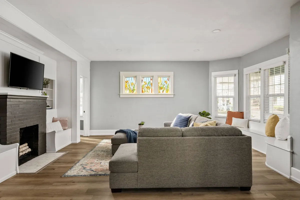 A warmly lit living room featuring a comfortable sofa, a classic fireplace with a glowing fire, stained glass window, and a television. The space is arranged for group relaxation and socializing, inviting guests to gather and unwind.