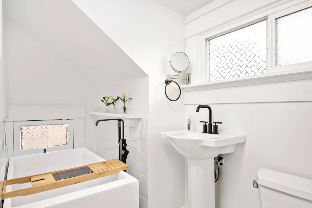 A bright, tranquil bathroom featuring a freestanding bathtub with a wooden bath tray and decorative flowers, a modern sink with a sleek faucet, and soft natural light filtering in. The design is both historic and updated, ideal for relaxing soaks.