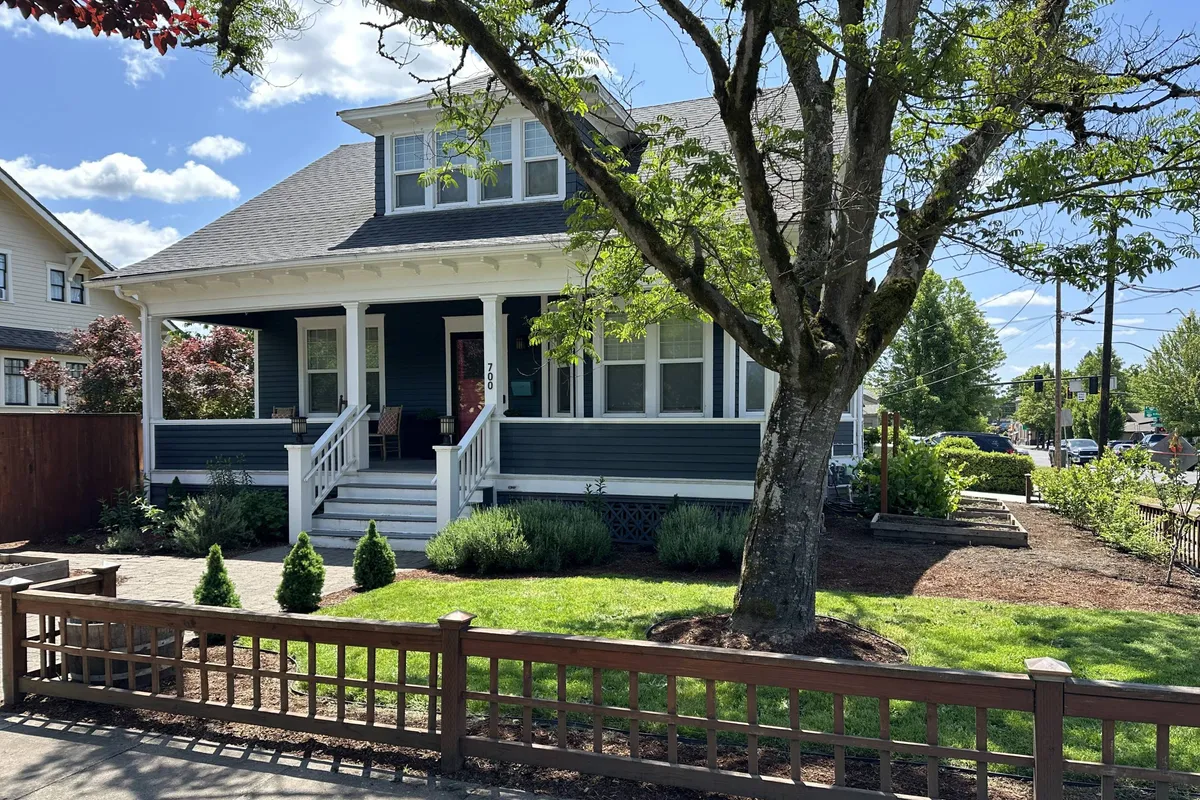 The image shows the inviting exterior of a single-family home with a welcoming front porch, steps leading up to the entrance, a large tree providing shade, and a manicured lawn bathed in soft natural light.