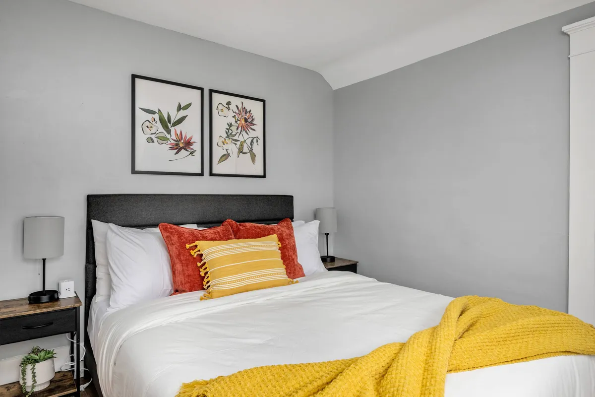 A comfortable bedroom featuring a bed with colorful pillows, framed floral art above the headboard, and nightstands with lamps on either side. The room is warmly lit, creating a cozy and inviting atmosphere, with plush bedding that promises restful nights.