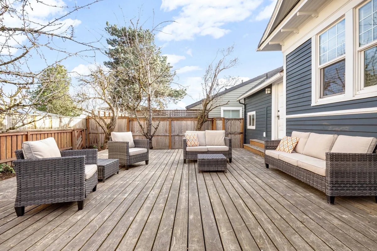 A serene outdoor seating arrangement on a wooden deck, accented with decorative pillows and surrounded by seasonal blooming flowers. The tranquil setting overlooks garden views bathed in warm sunset light, ideal for relaxing or socializing.