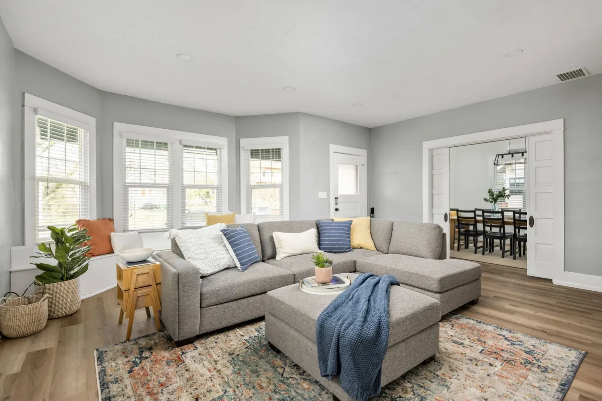 A cozy living room featuring a sectional sofa atop a decorative rug, accented by a leafy plant. Through a wide doorway, the dining area is visible, suggesting an open-concept flow between spaces and inviting opportunities for relaxation or social gatherings.