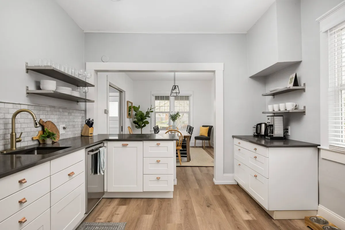 A modern, sunlit kitchen and dining space featuring a kitchen island, open shelving, and natural light streaming through large windows. The setup encourages collaborative cooking and casual gatherings.