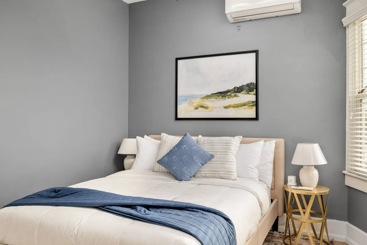 A serene bedroom scene with a neatly made bed, a decorative pillow arrangement, a soft lamp glow, and a tasteful painting above the headboard. The overall mood is quiet and calming, echoing the blend of historic charm and modern touches.