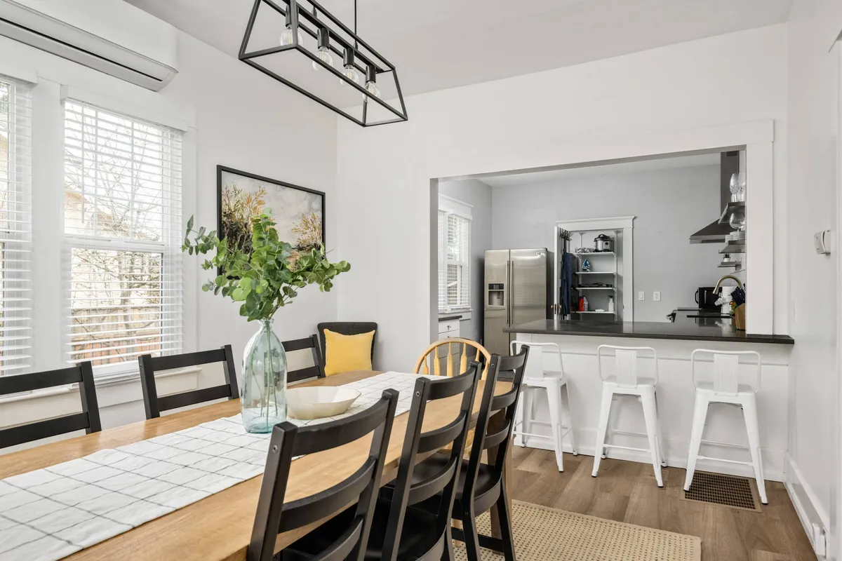 An inviting dining area with a large table, greenery in a vase, stylish artwork, and bar stools at the kitchen counter. The space exudes a welcoming mood ideal for group meals and celebrations.