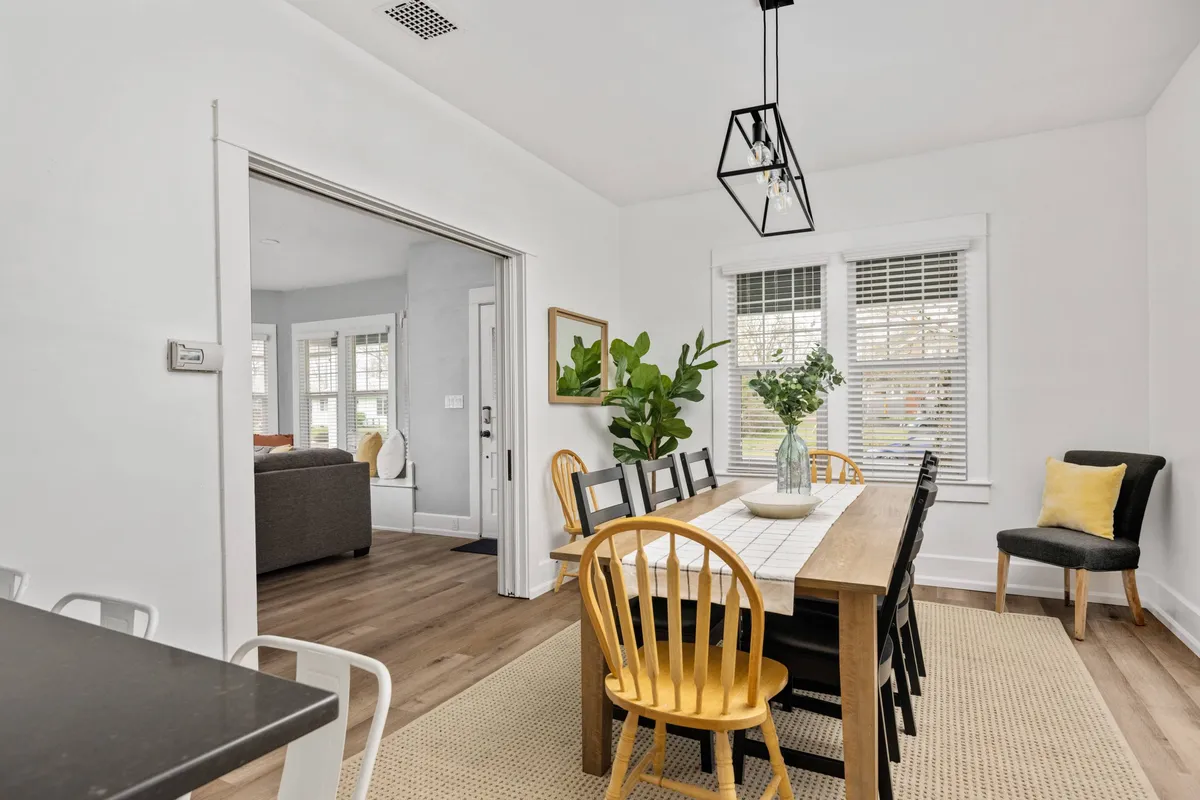 A stylish dining area with a wooden table, comfortable chairs, a leafy plant centerpiece, and a standout lighting fixture above. The setting is intimate, bathed in warm light, and ready for meals or lively conversation.