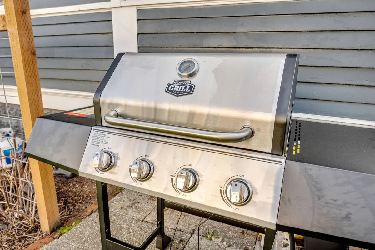 A close-up exterior shot featuring a stainless steel BBQ grill with visible control knobs and a temperature gauge, set up for outdoor cooking. The scene is ready for grilling, evoking images of social outdoor dining and family barbecues.